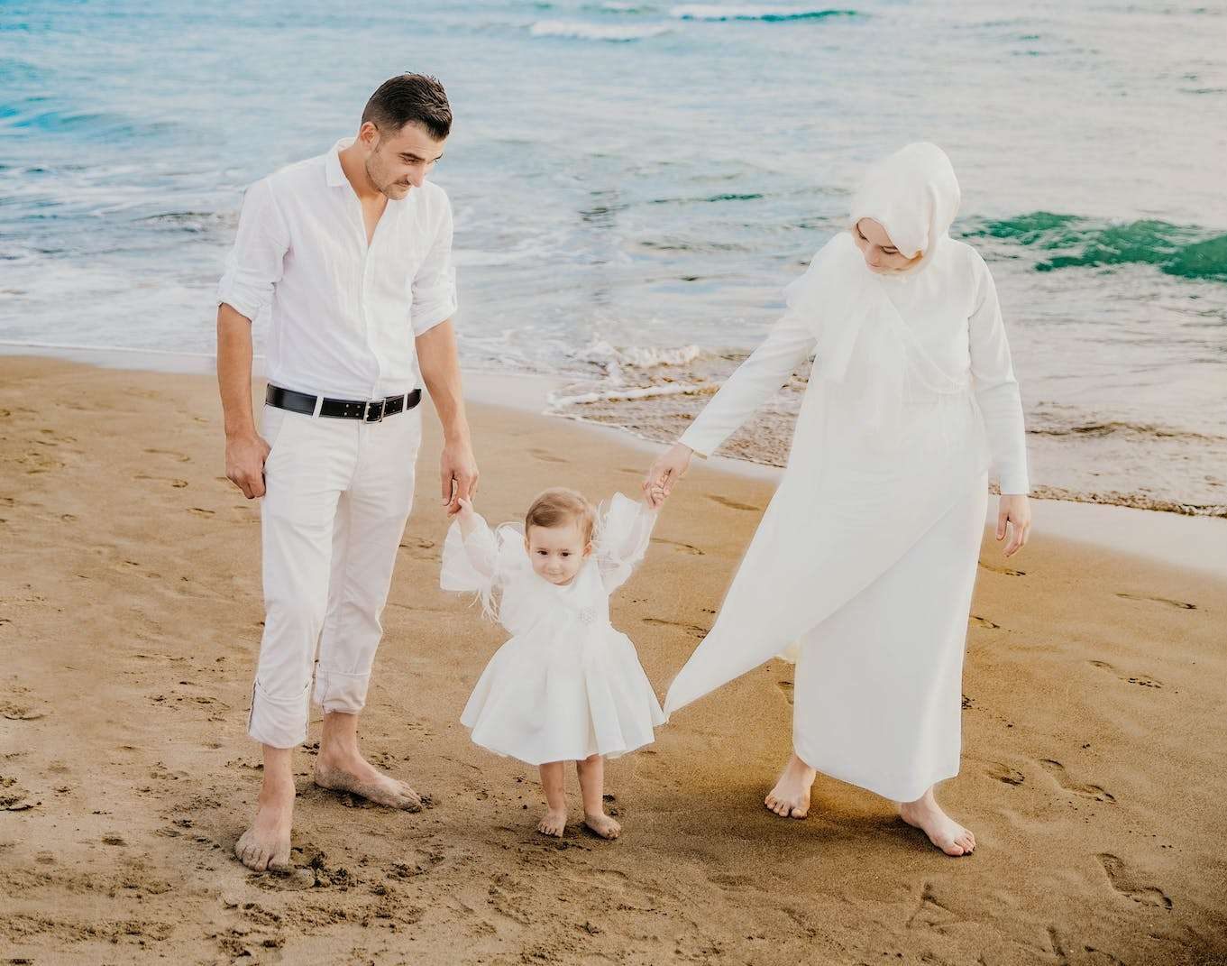 Full length of Muslim couple in stylish white clothes holding hands of little daughter while walking on sandy coast near wavy ocean in sunny day